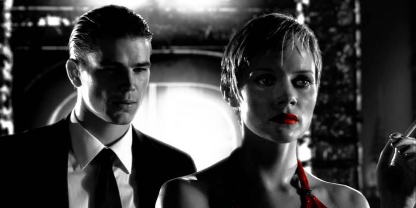 A man in a suit stands behind a woman on a balcony, her red dress and lipstick strikingly red against the black and white aesthetic.