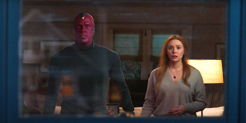 Vision and Wanda stand next to one another looking out the living room window, worried, in WandaVision