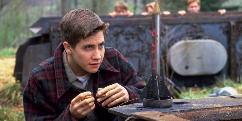 Jake Gyllenhaal crouches down outdoors by a small rocket in October Sky.