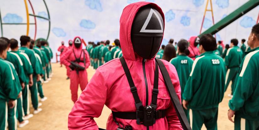 Contestants in matching jumpsuits line up while masked officials walk by with weapons in Squid Game