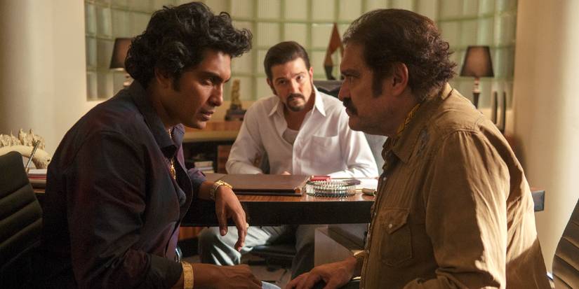 Diego Luna watches Tenoch Huerta talk to an older man from behind his desk in Narcos Mexico
