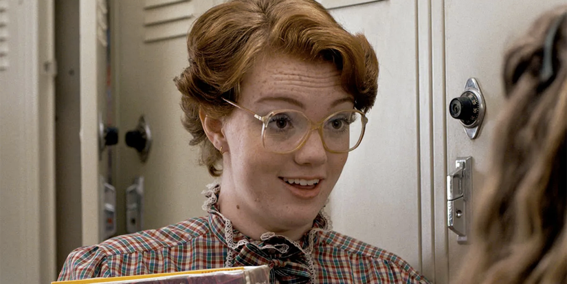 Shannon Purser as Barb Holland with glasses talking in a school hall in Season 1 of Stranger Things.