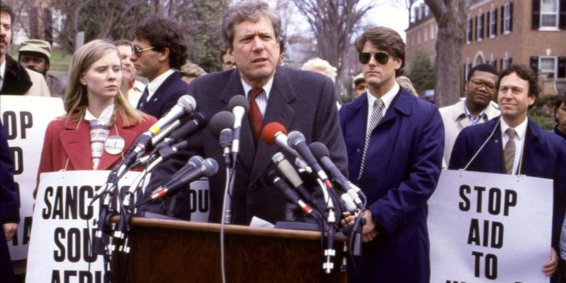 A man talks at a podium with many microphones as people hold signs behind in Tanner '88.