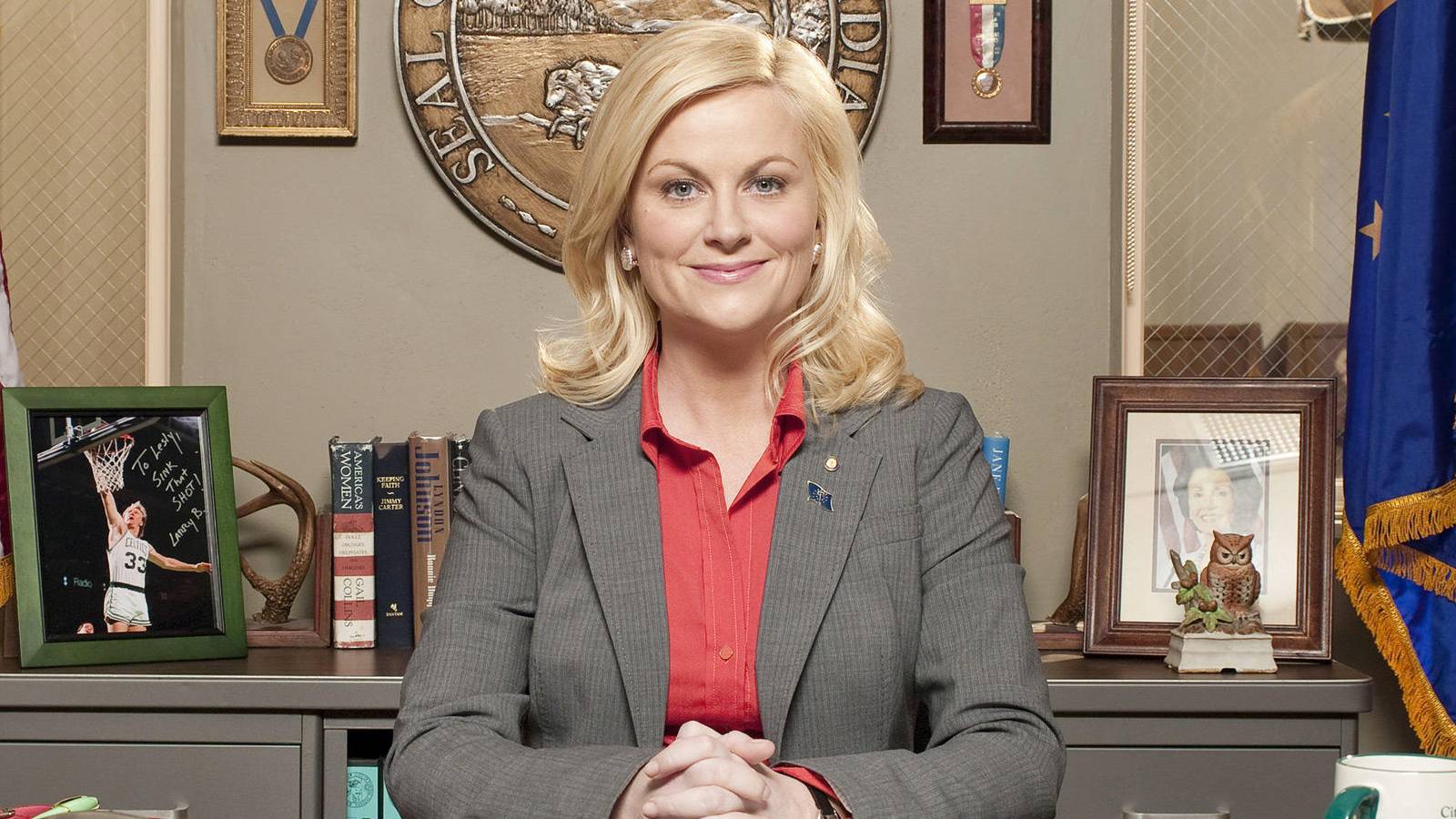Amy Poehler as Leslie Knope sitting behind her desk and smiling for the camera in Parks and Recreation.