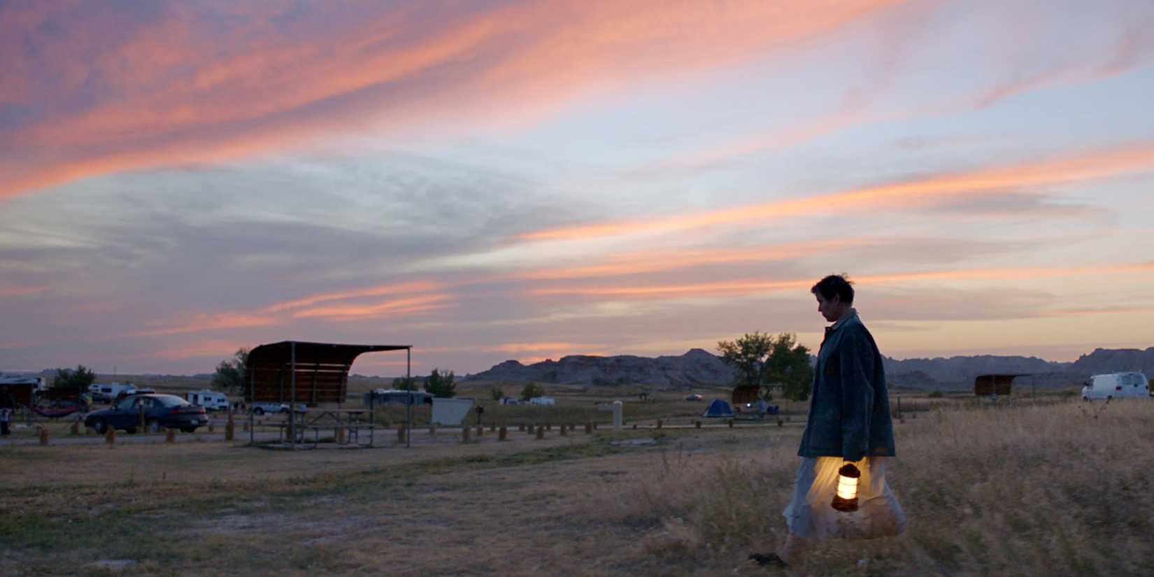 Frances McDormand walking through a field with a lamp, in front of a sunset in Nomadland.