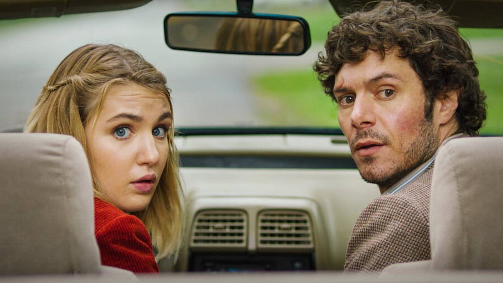 Sophie Nélisse and Adam Brody looking behind over their shoulders as they sit in a car in The Kid Detective.