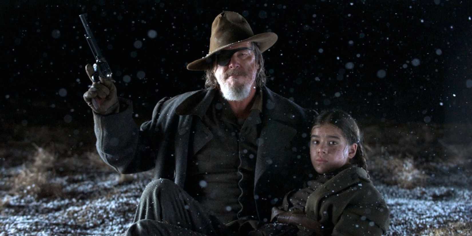 Rooster Cogburn (Jeff Bridges) kneels beside a wounded Mattie Ross (Hailee Steinfeld), aiming his gun up into the snowy night in True Grit