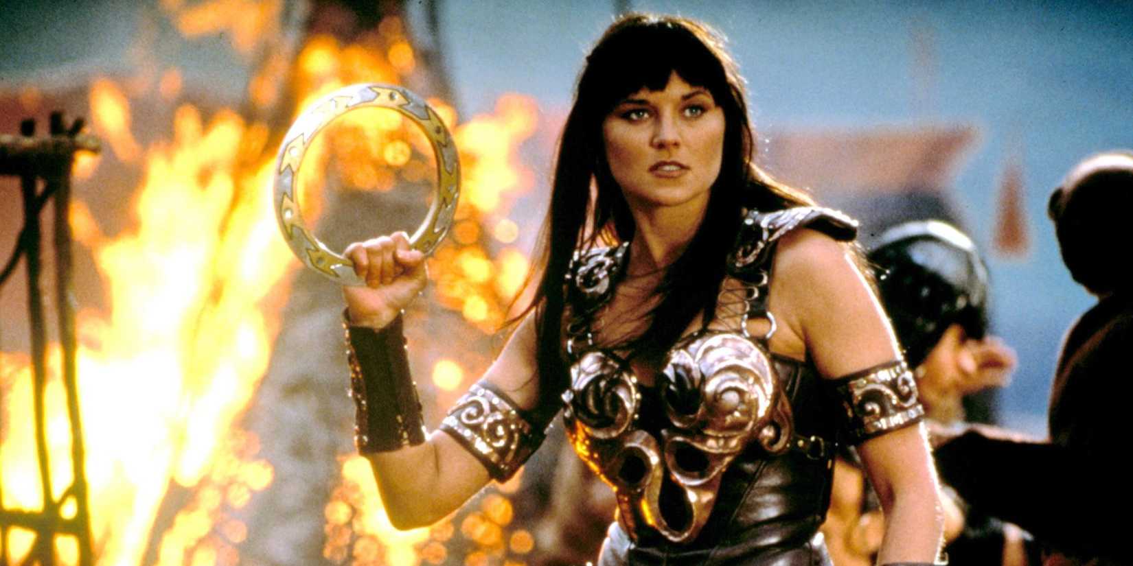 Lucy Lawless as Xena holding a weapon with a burning building in the background in Xena: Warrior Princess.