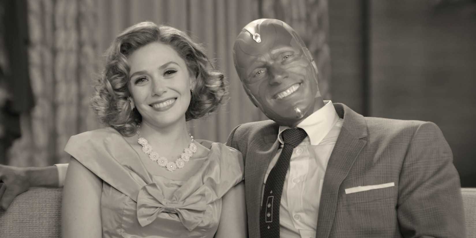 A woman and a robot-like man smile at the camera while sitting on the sofa in WandaVision.