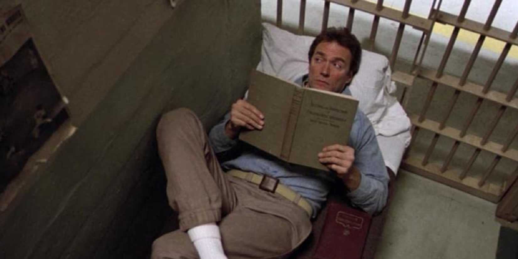 A man reads a book lying on a bed in a prison cell in Escape From Alcatraz.