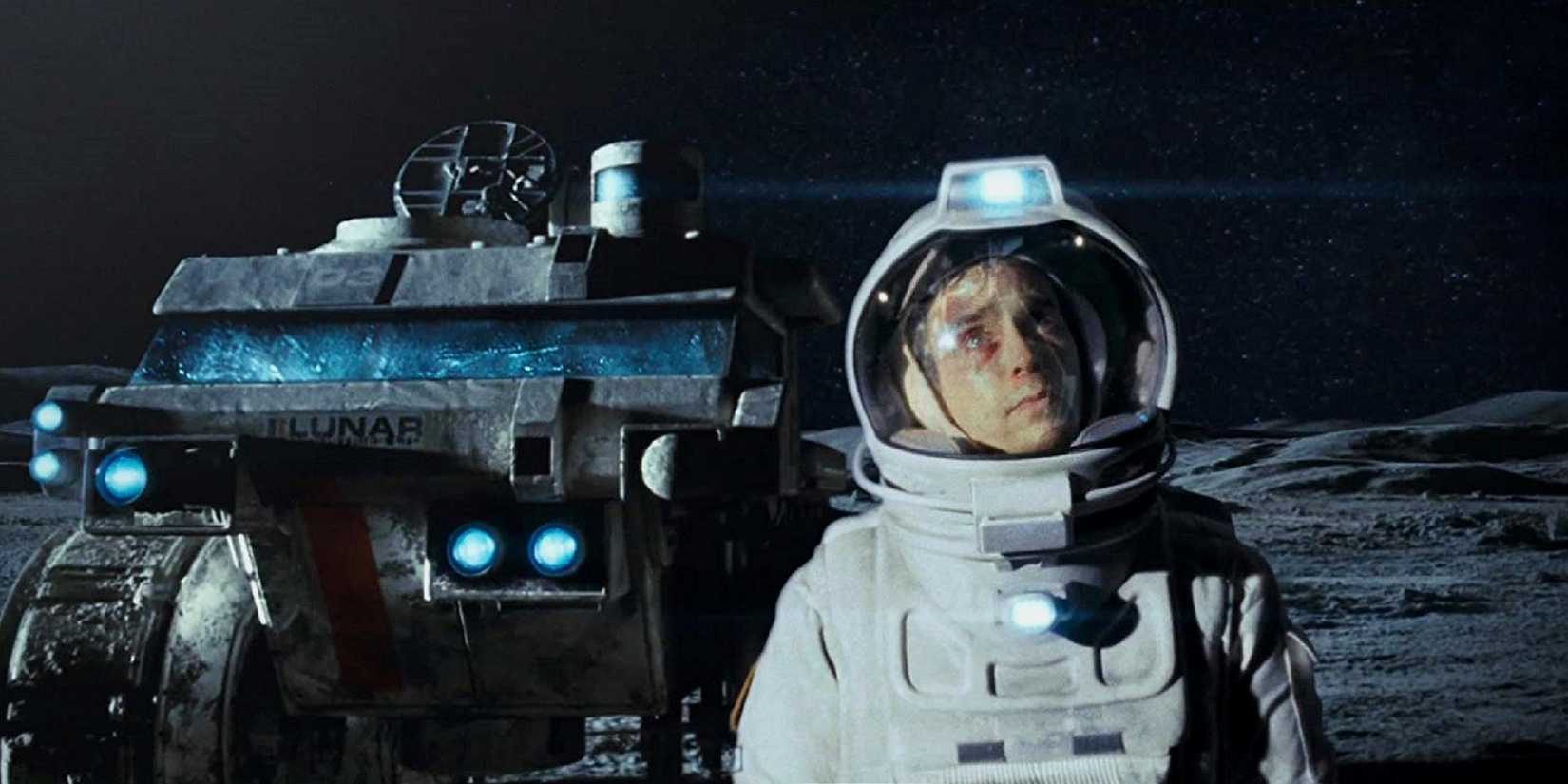 Sam Rockwell in a white spacesuit standing on the moon's surface, with a gray mining vehicle behind him, looking up at the stars, in Moon
