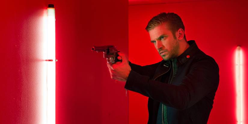 Dan Stevens stands in a red room pointing a gun