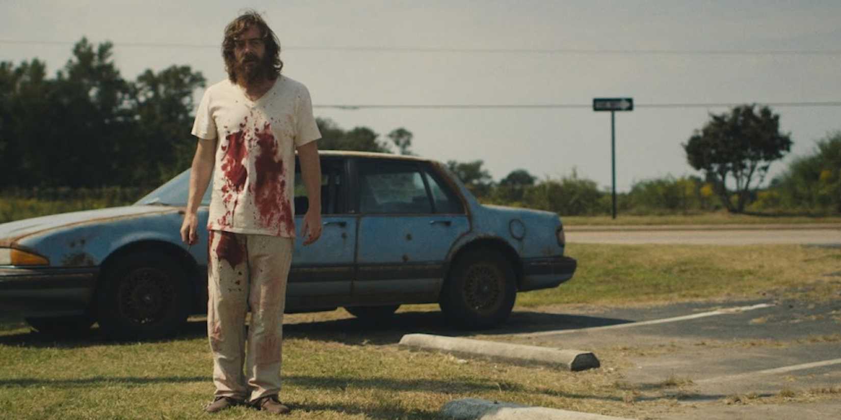 A man with blood on his shirt standing beside a car in Blue Ruin.
