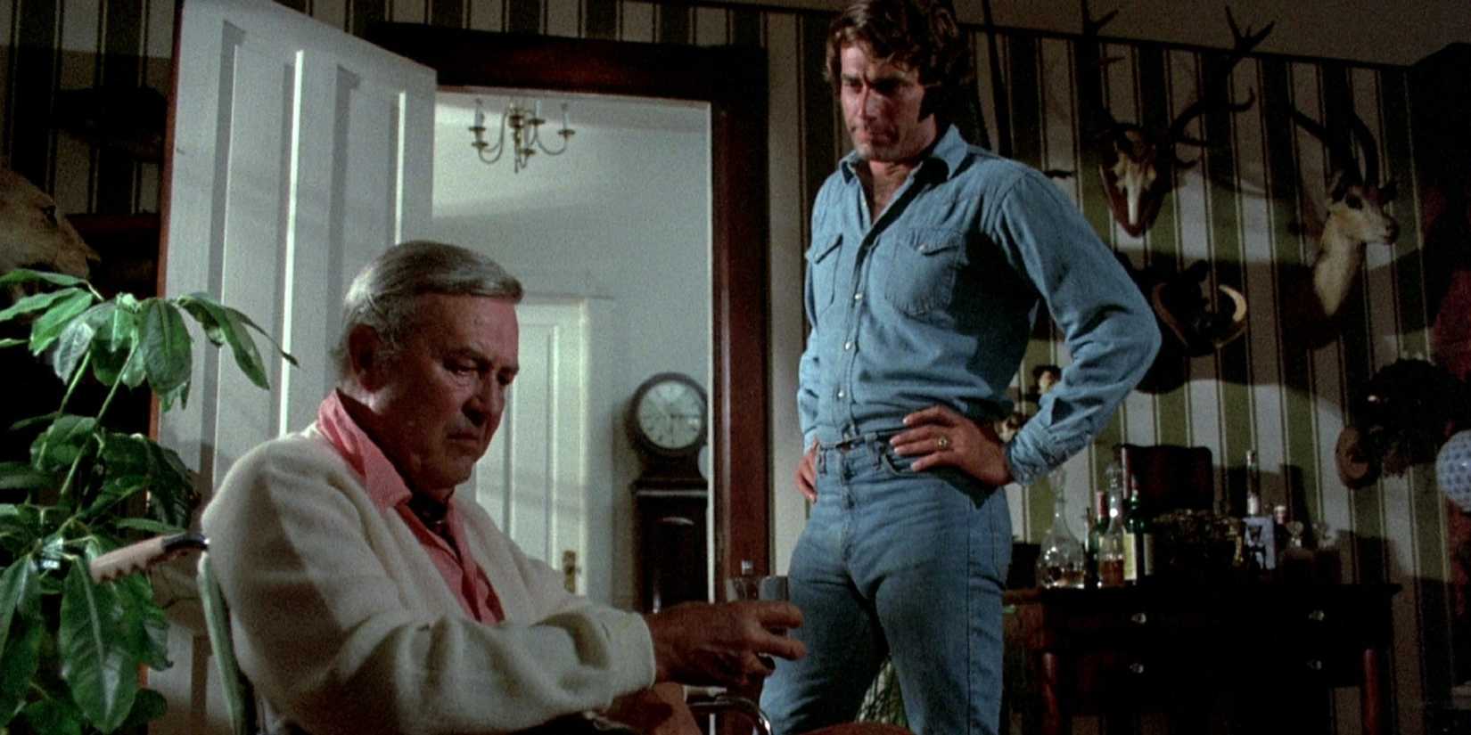 A man stands next to another man sitting on a chair
