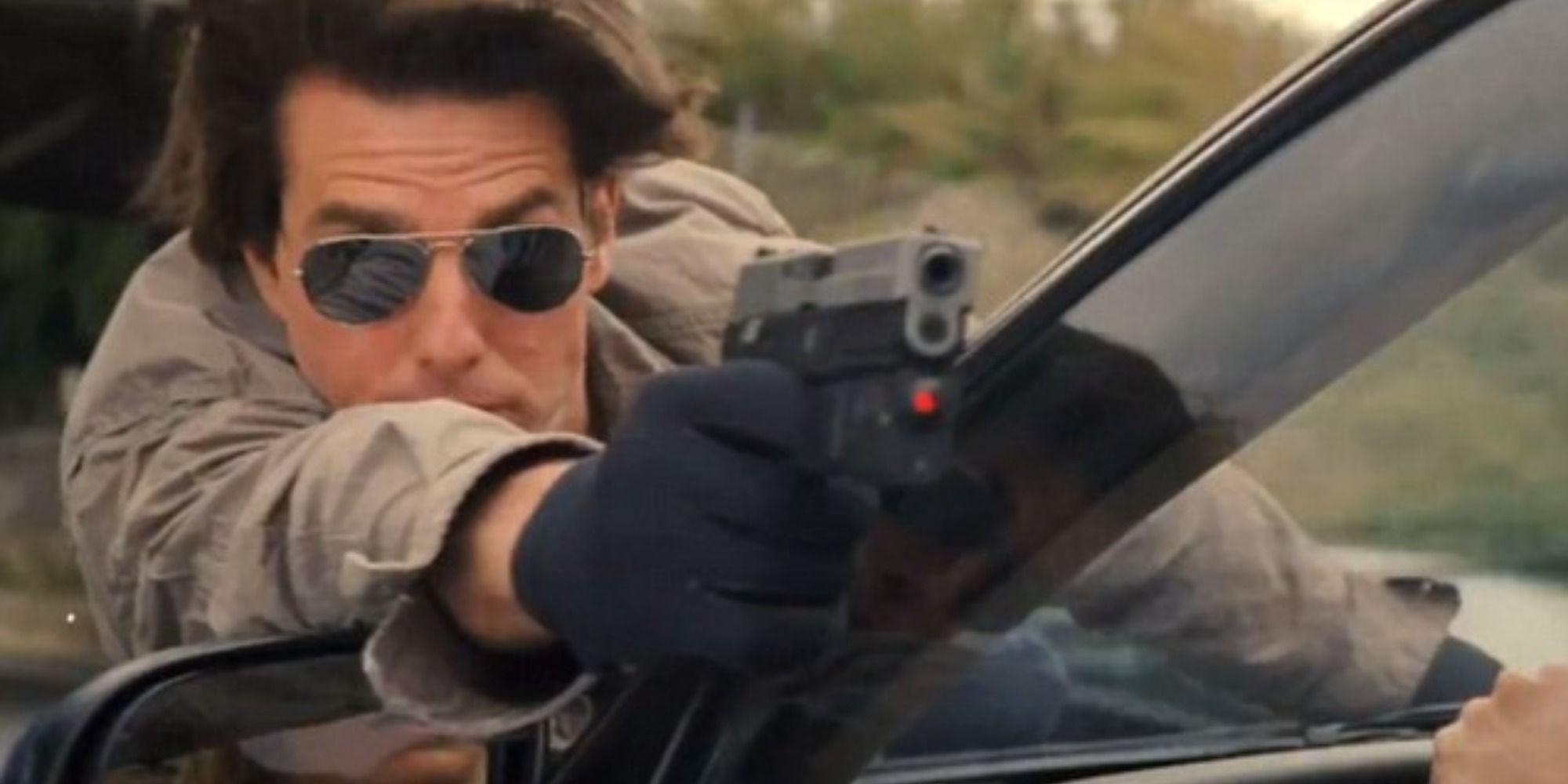 Tom Cruise hanging on the front of the car while aiming a gun 