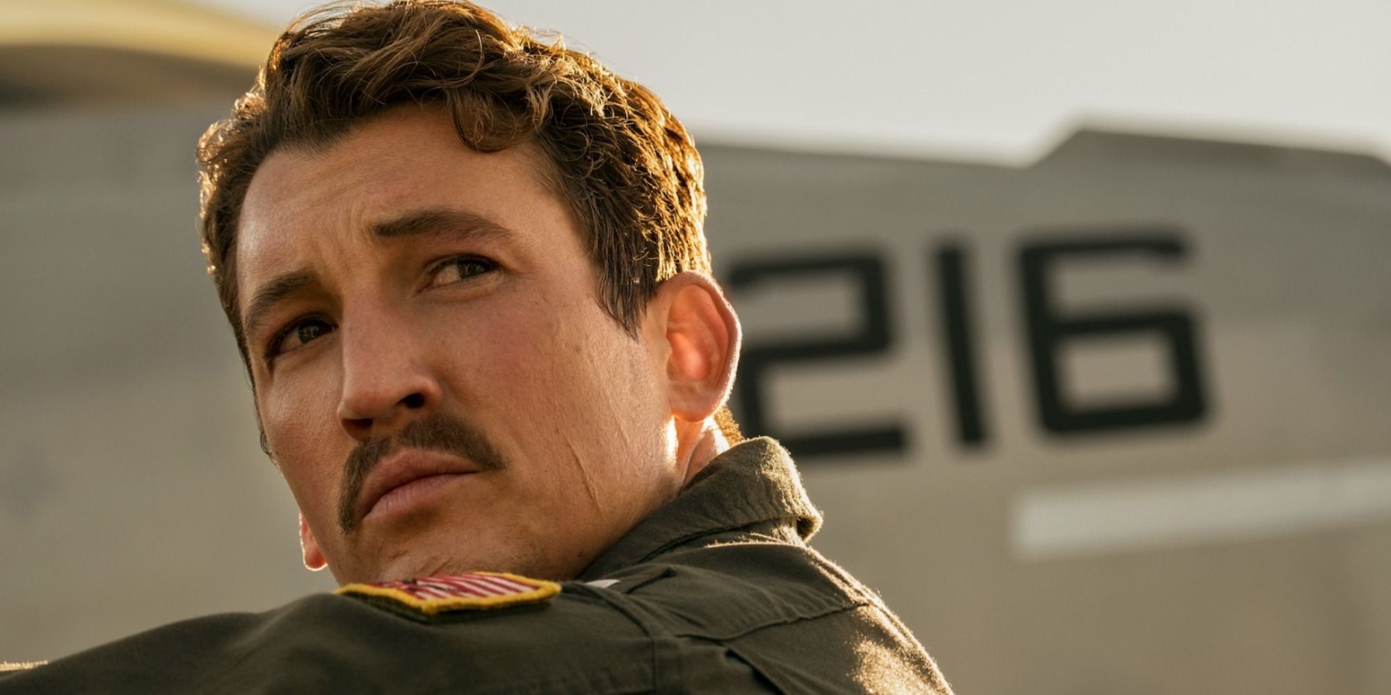 Miles Teller as Rooster beside a plane looking off to the side 