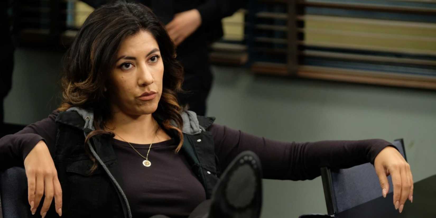 Stephanie Beatriz as Rosa Diaz sitting in a chair in the office looking unamused in Brooklyn Nine-Nine.