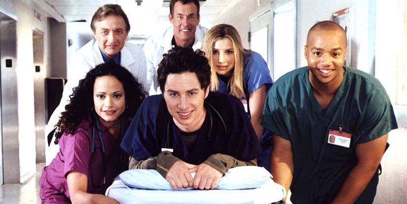 The cast of Scrubs face the camera and smile on the set, inside a hospital in a promo photo. 