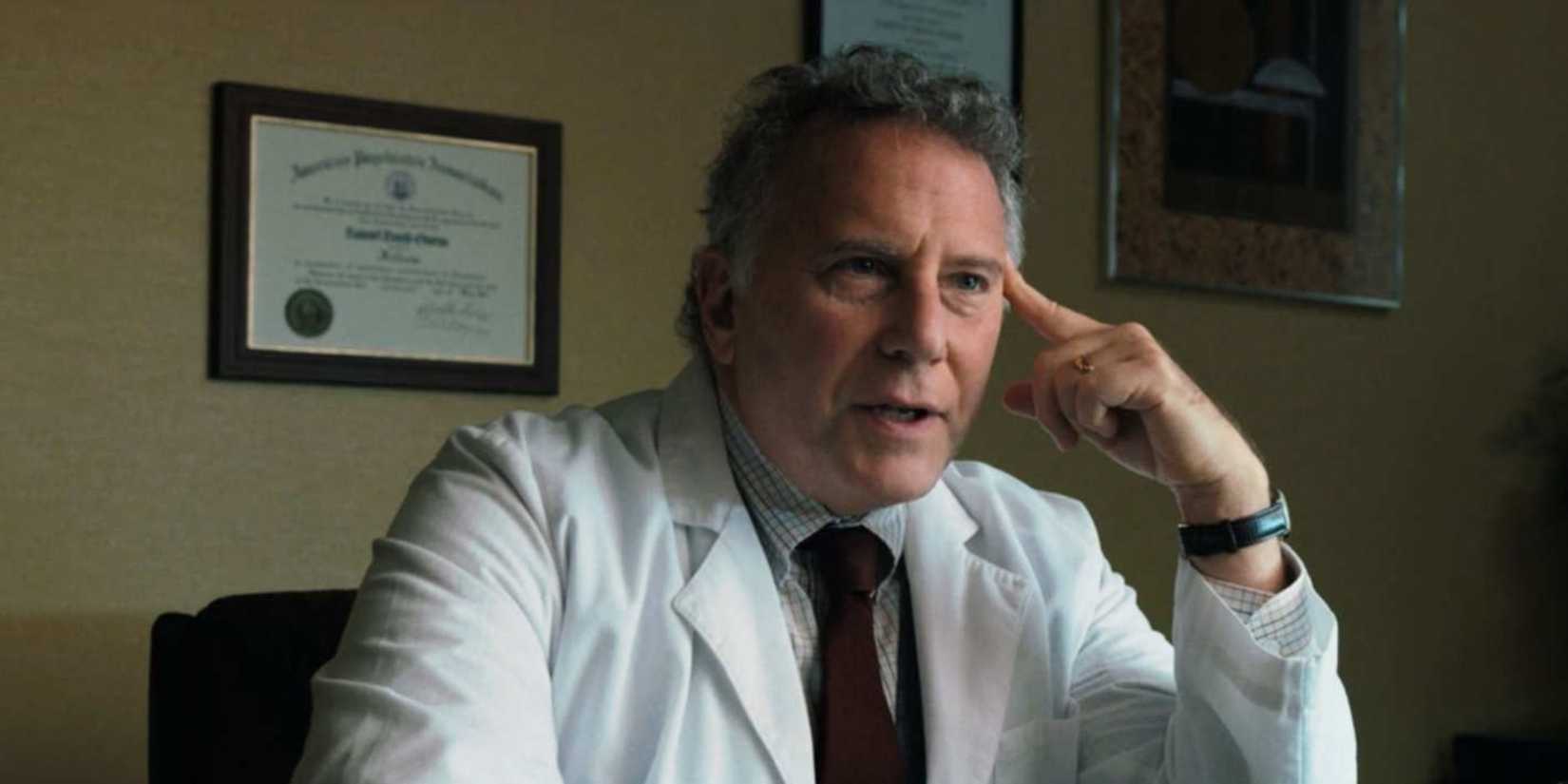 Paul Reiser as Dr. Sam Owens in a white lab coat and tie is sitting at his office desk in Stranger Things.