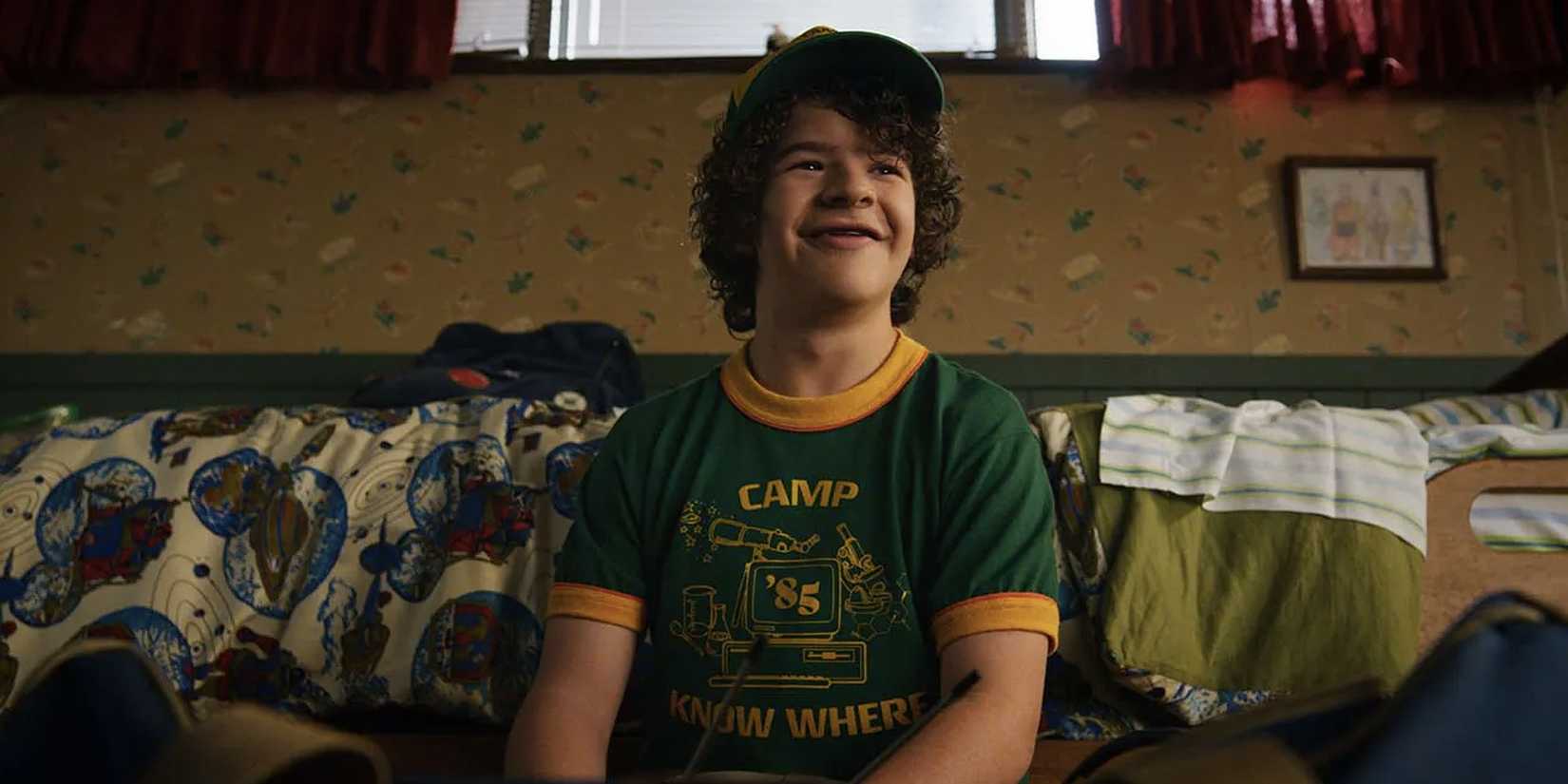 Dustin sits on a couch with a "Camp Know Where" shirt on and a smile on his face in 'Stranger Things'.