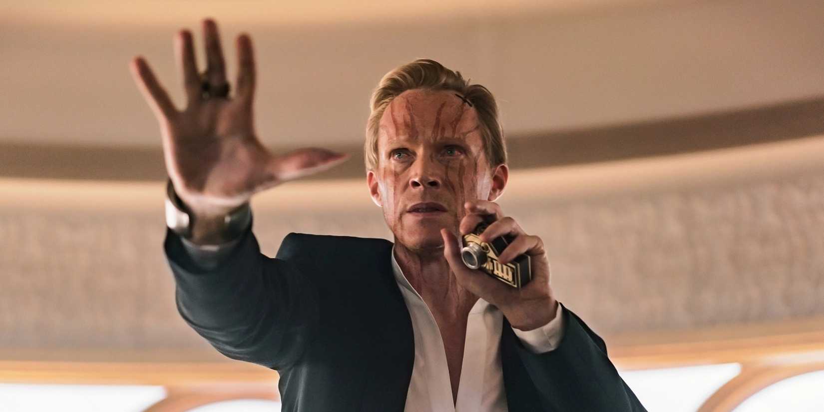 Dryden Vos, wearing a suit with a scarred face, stands with his right hand outstretched in front of him