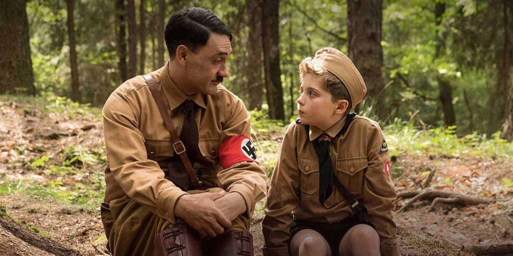 Taika Waititi as Imaginary Adolf Hitler sitting beside Roman Griffin Davis as Jojo in the forest in Jojo Rabbit