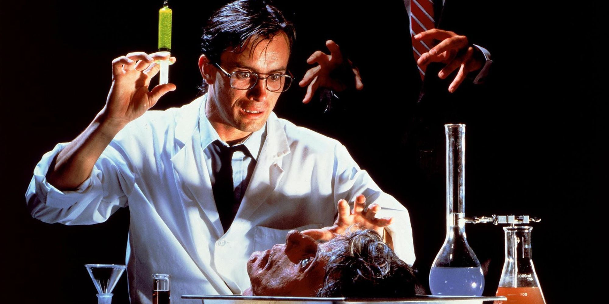A man in a lab coat looks down at a human head on a table with scientific jars nearby in Re-animator.