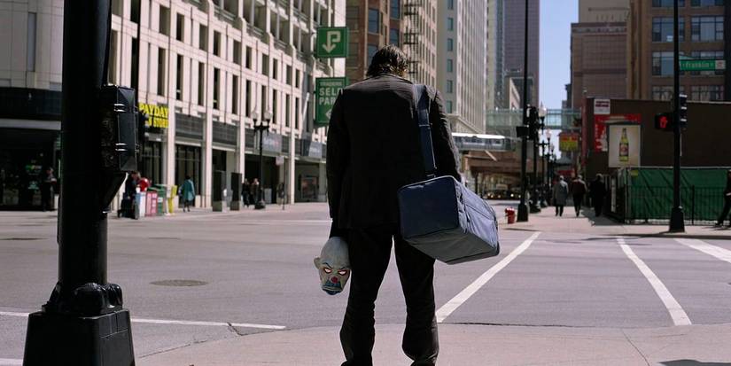 A shady man with a mask and a duffle bag stands in the city streets. 
