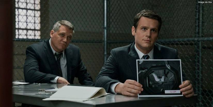 FBI agent Holden Ford (Jonathan Groff) holds a black and white photograph and sits next to FBI agent Bill Tench (Holt McCallany) in Mindhunter.