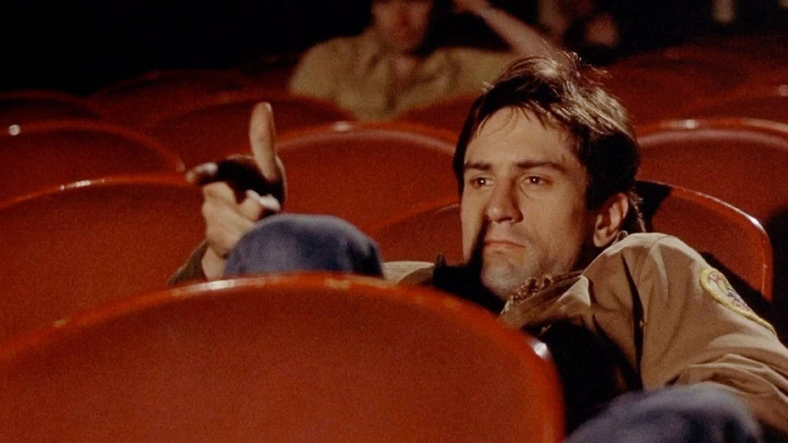Travis Bickle pointing a finger gun at the movie screen while sitting in a red movie theater seat in 'Taxi Driver.'
