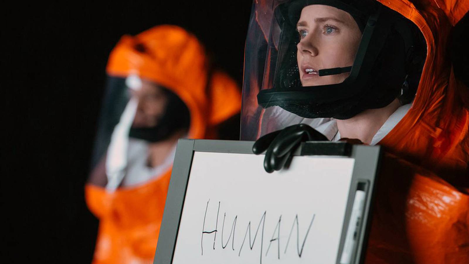 Louise Banks wearing a hazmat suit and holding a whiteboard that says 