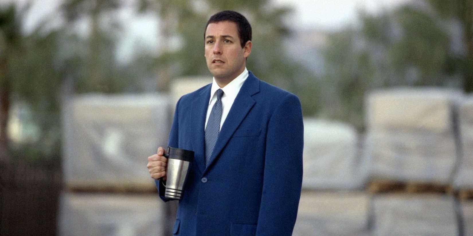 Adam Sandler as Barry anxiously standing with his thermos outdoors in front of pallets in Punch Drunk Love.