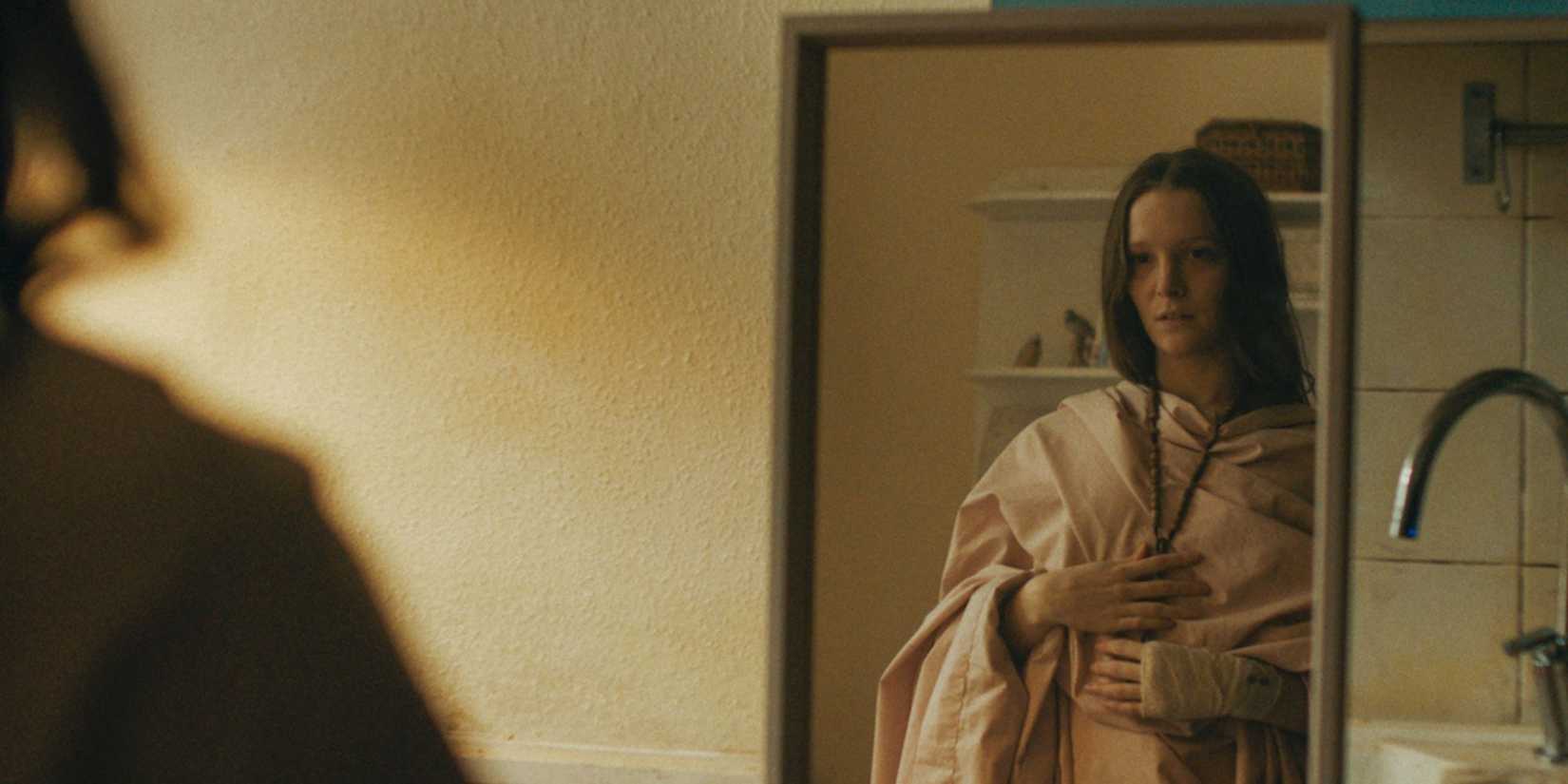 A woman dresses herself in a robe and stands in front of a mirror