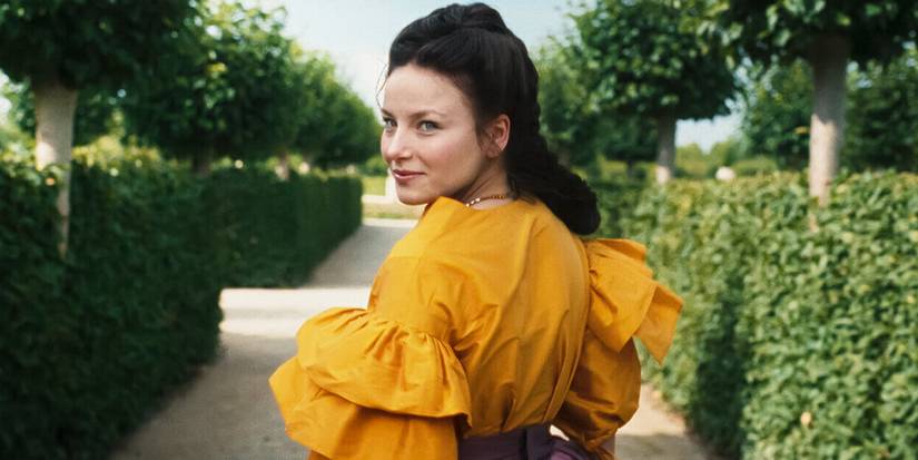 Devrim Lingnau wearing a yellow period dress and standing in the middle of a garden in 'The Empress'