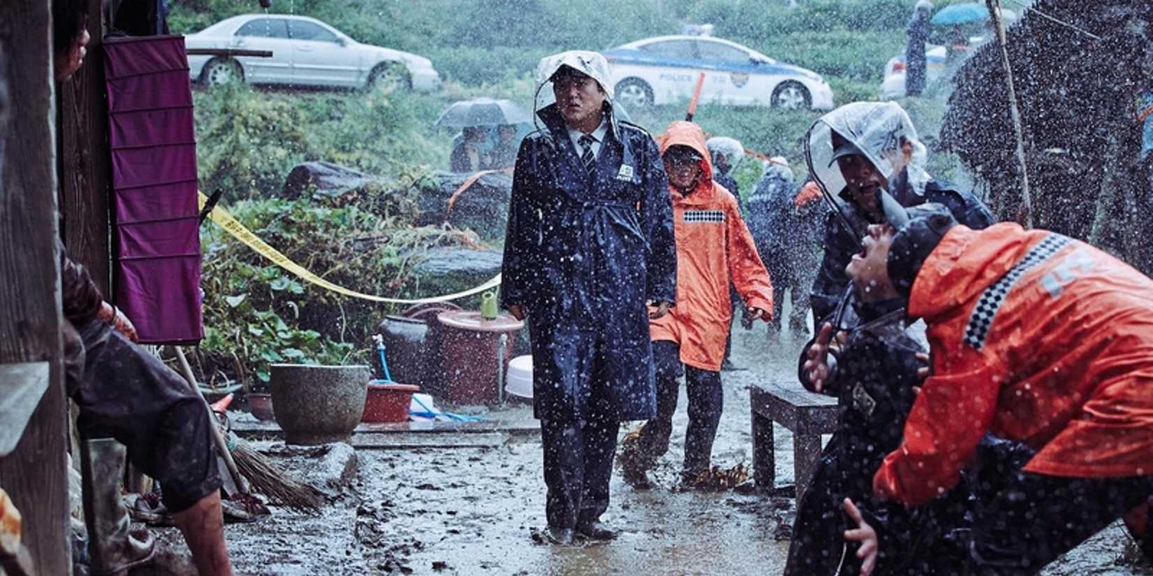 People in rain gear walk through a muddy street with caution tape in the background in The Wailing (2016).