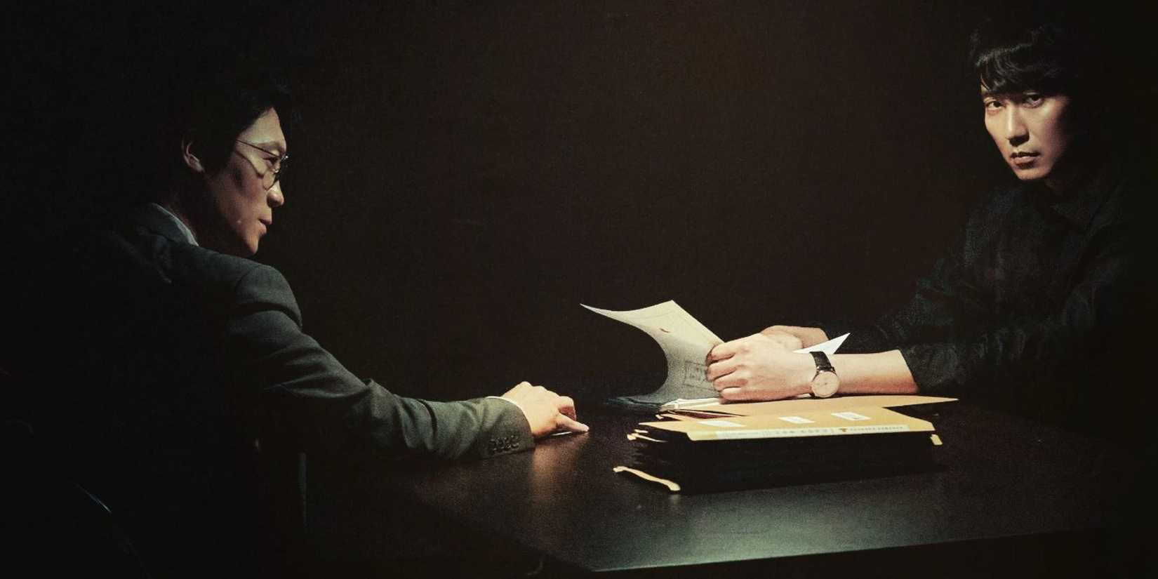 A scene from Through the Darkness where two men sit at a table in darkness and one has a stack of files.