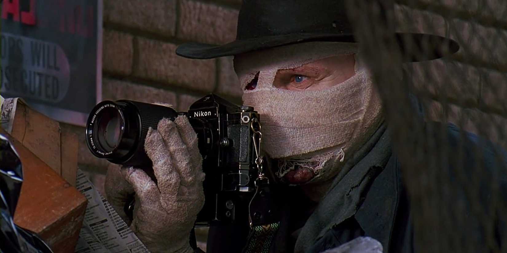 A masked Liam Neeson with a hat taking photos from behind a net in Sam Raimi's Darkman