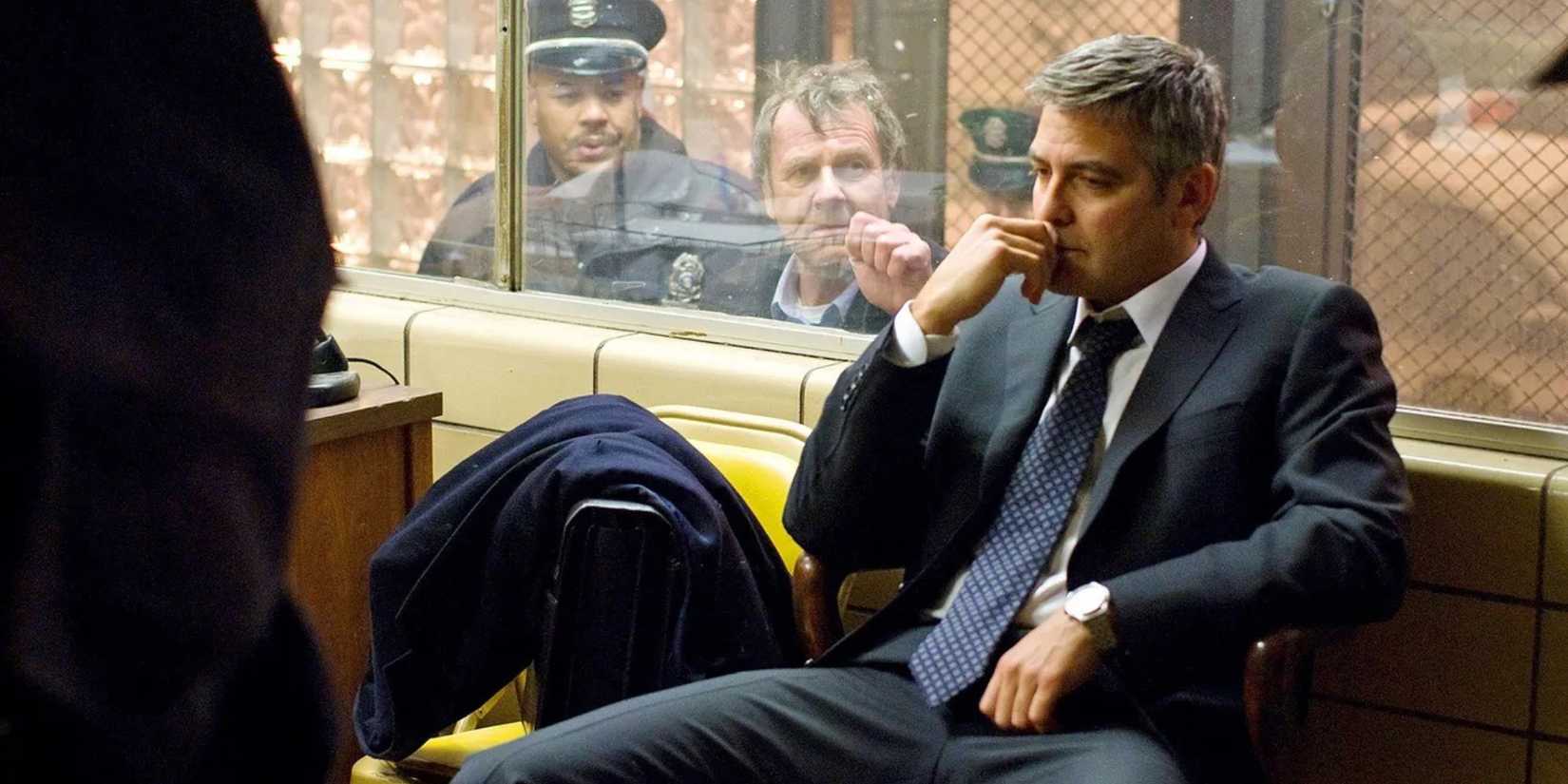 George Clooney in a police station looks disappointed as someone behind taps on glass in Michael Clayton.