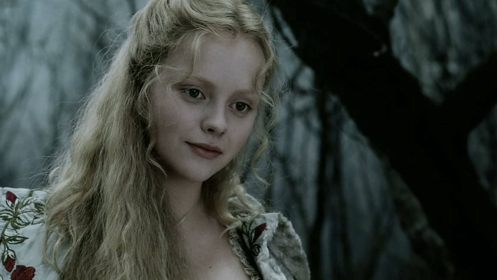 A pale blonde woman in Victorian era clothing stands in a dark forest