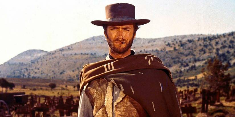 A cowboy in a poncho scowls as he smokes a cigarette while standing against a barren, sun-scorched backdrop.