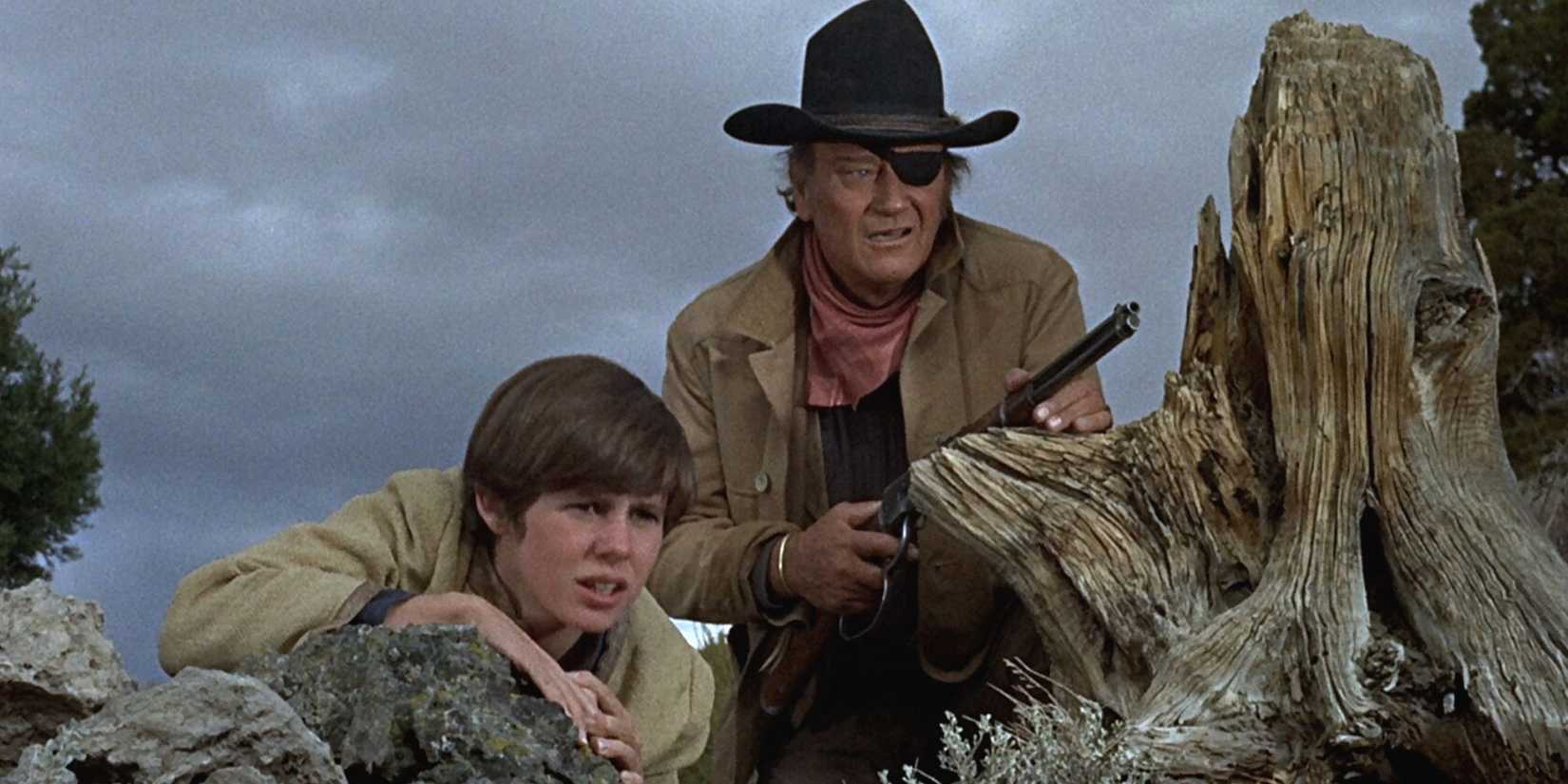 Kim Darby and John Wayne with a shotgun crouching down behind rocks looking out at something in True Grit.