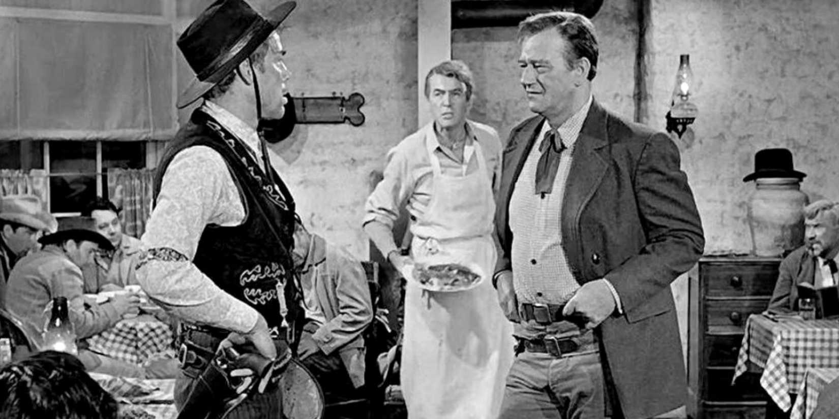 A cowboy and a gunslinger argue in a diner with their hands on their guns as a waiter behind them watches.