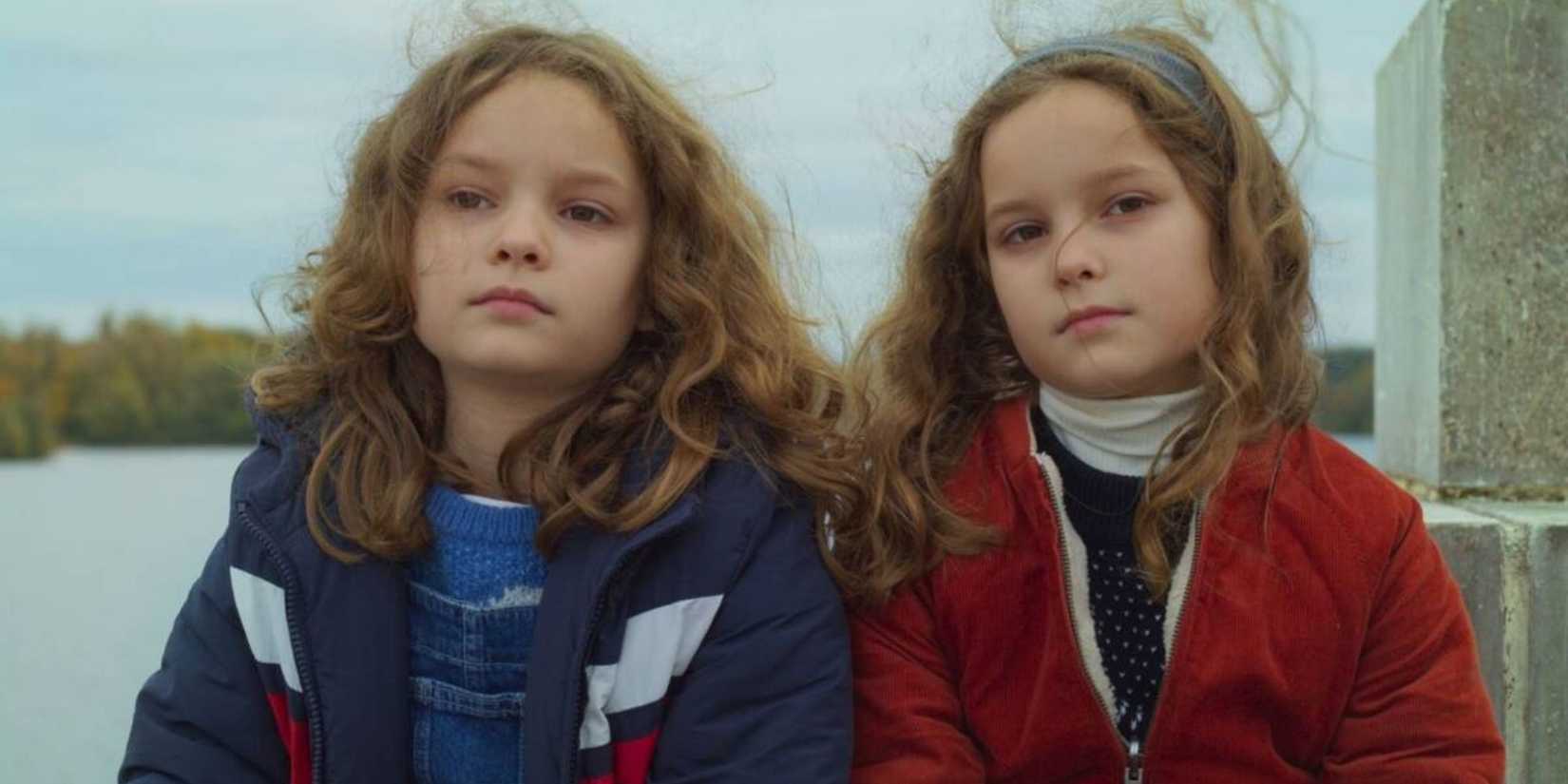 Two identical young girls sitting next to each other outdoors by a lake in Petit Maman.