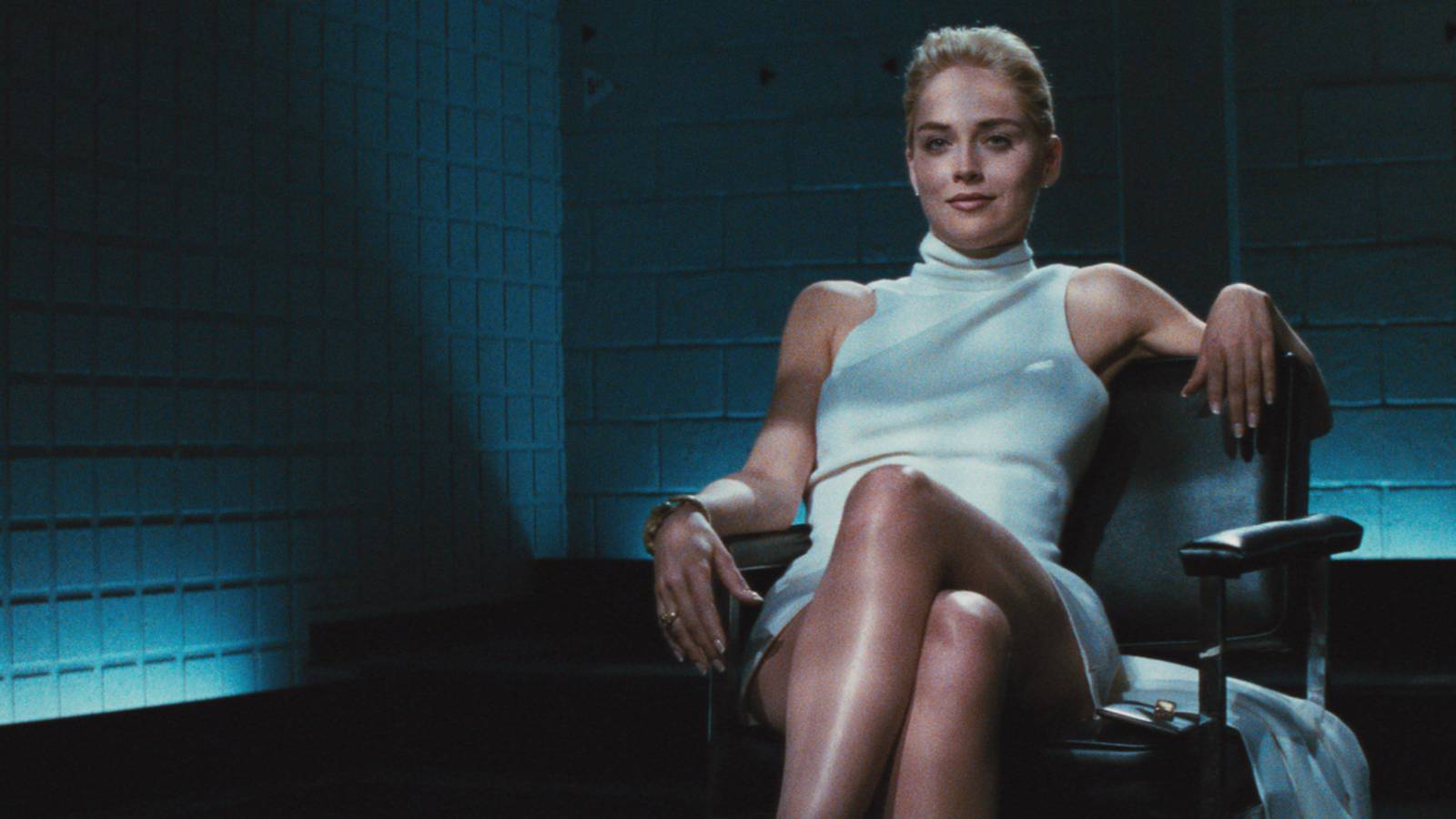 Sharon Stone as Catherine Tramell, seated in a chair in a white dress with her legs folded, in Basic Instinct.