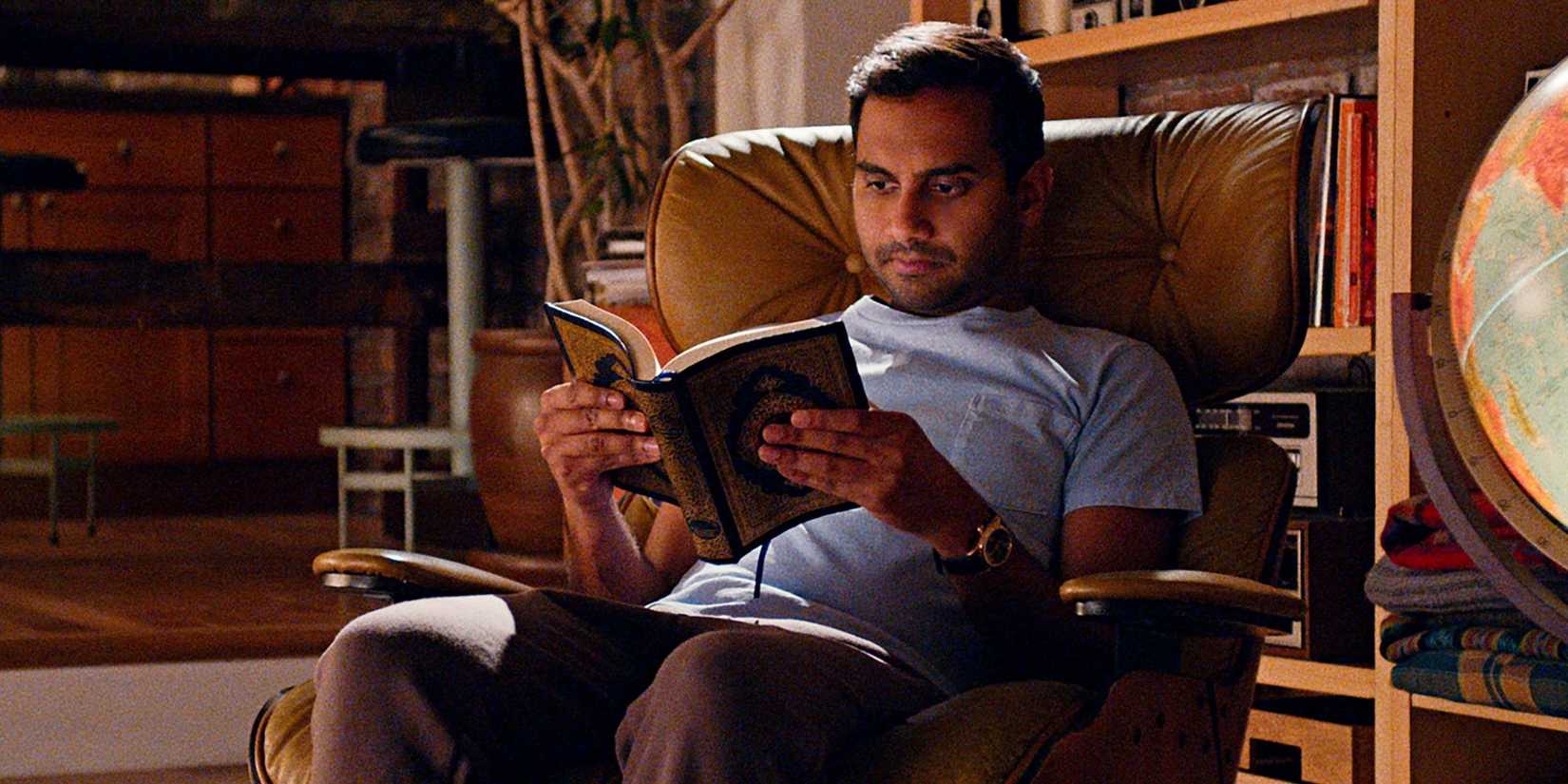 Dev from Master of None sitting in a recliner in the dark reading a book.