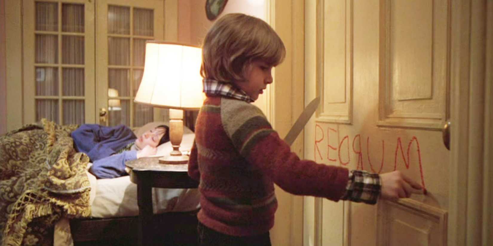 Danny Lloyd as Danny Torrance writing redrum on door holding knife: mother sleeping in bed in background