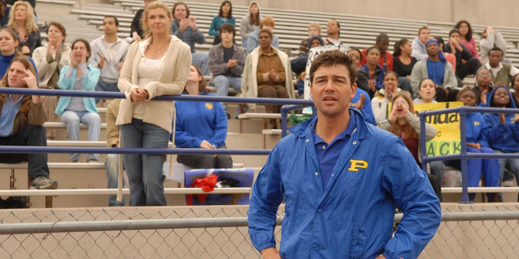 Coach Eric Taylor stands on the football field sidelines while his wife, Tami, stands in the bleachers behind him. 