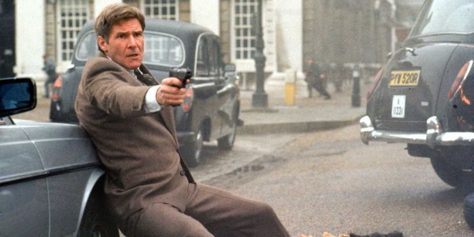 Harrison Ford as Jack Ryan leans against a car and aims his gun at someone off-camera in 'Patriot Games'.