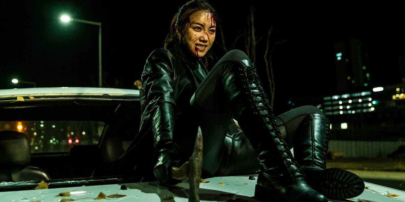 A bloodied, weary assassin sits upon the hood of a damaged car. 