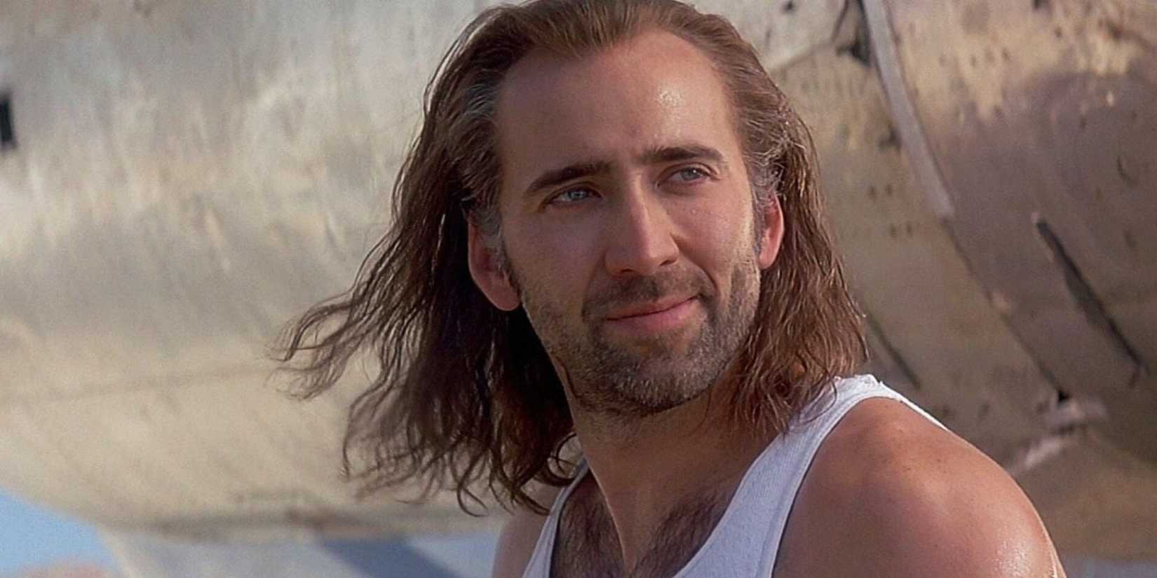 Nicolas Cage with long hair stands in front of an airplane and smiles and looks off camera in Con Air.